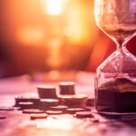 Sand running through an hourglass next to small stacks of coins against a setting sun