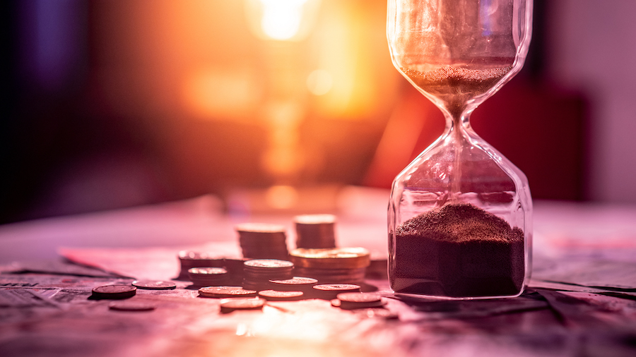 Sand running through an hourglass next to small stacks of coins against a setting sun