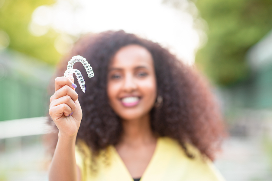 Smiling Black woman wearing a yellow blouse is blurred in the background as she holds up 2 Invisalign aligners outside