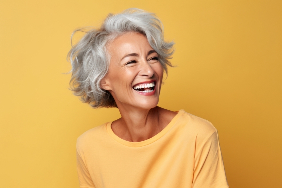 Older woman with a CEREC dental crown smiles brightly while wearing a yellow blouse against a yellow wall